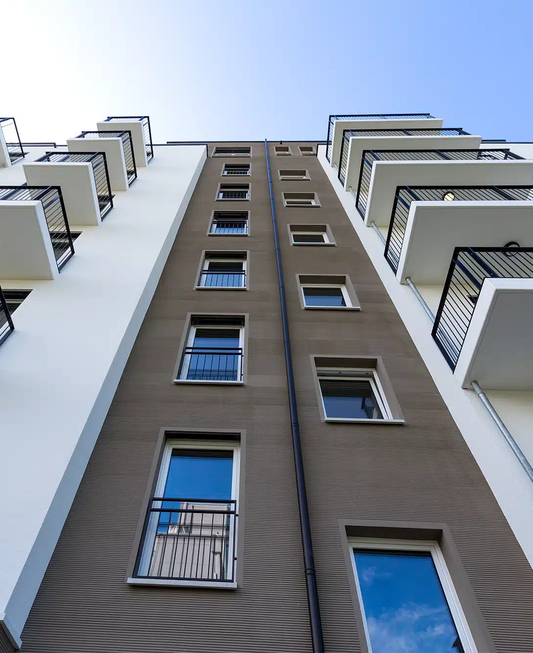 Gebäude aus Froschperspektive fotografiert. Fassade in der Mitte in nBraun mit leichten Streifen. Rechts und Links Weiße Fassade mit schwarzen Balkonen